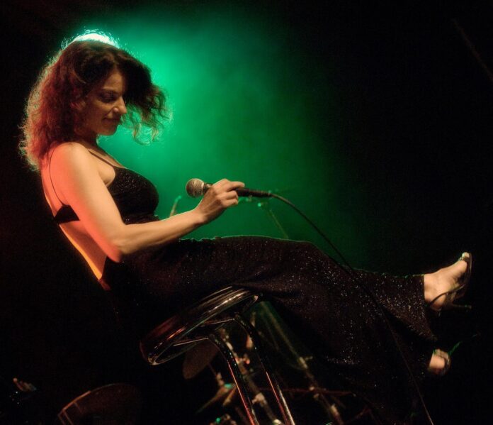 Cantante jazz seduta sul palco durante una performance con microfono in mano e luci colorate sullo sfondo.