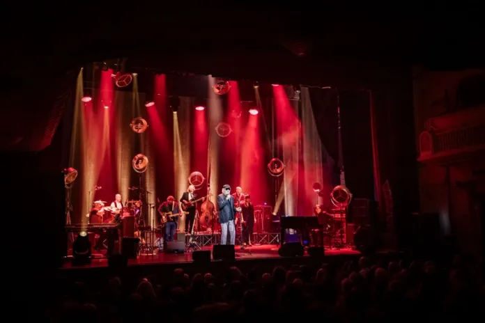 Veduta ampia di un palco illuminato di rosso, con un cantante al centro, musicisti e pubblico in primo piano.