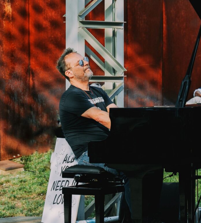 Pianista seduto al pianoforte in un ambiente esterno con struttura di metallo visibile sullo sfondo.