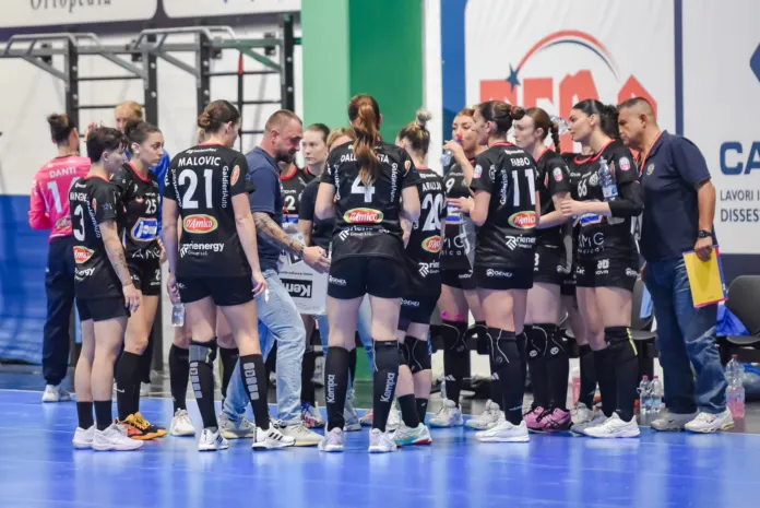 Fotografia di una squadra femminile di pallamano durante un momento di pausa o discussione, in un'area sportiva indoor.
