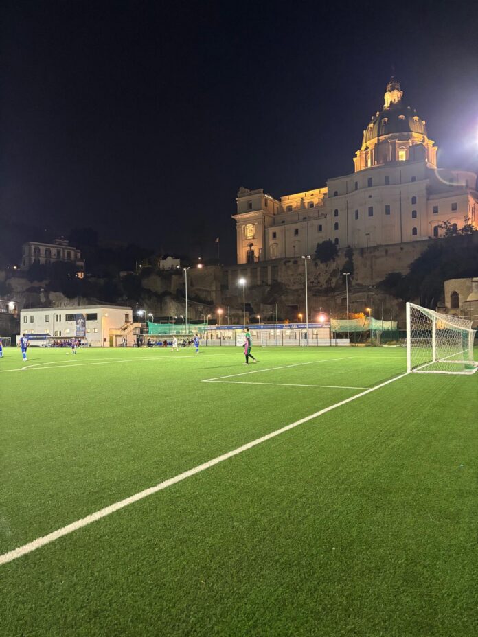 Campo da calcio di sera con luci accese e edificio monumentale illuminato sullo sfondo.