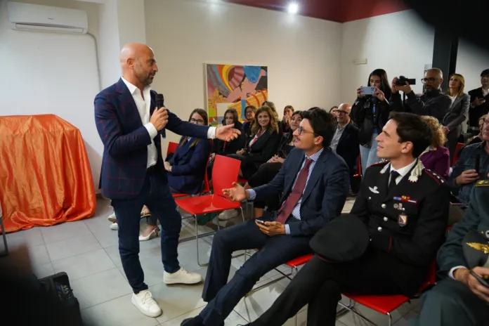 Un uomo in piedi con un microfono si rivolge a persone sedute; nella sala sono presenti altre persone in piedi.