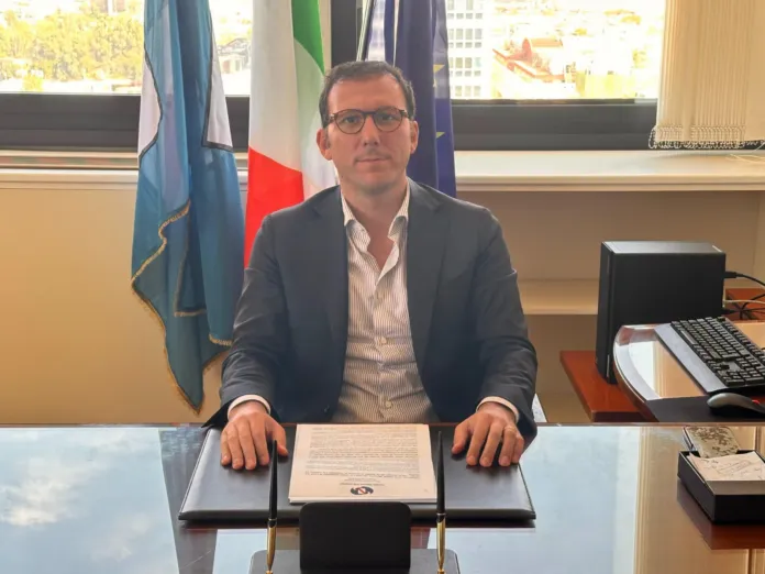 Consigliere regionale Giovanni Maria Cuofano seduto a una scrivania in ufficio con bandiere sullo sfondo.