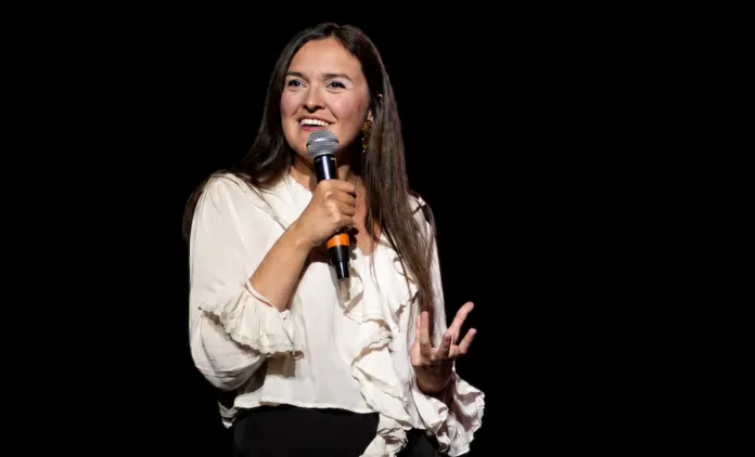 Maura Bloom con microfono in mano mentre parla sul palco con sfondo nero