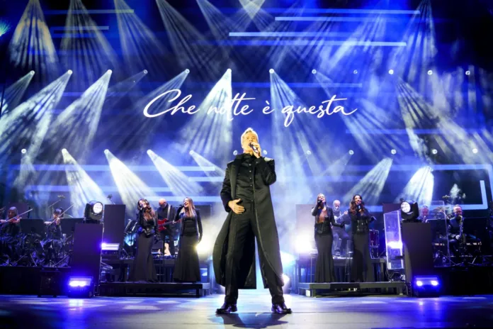 Un cantante al centro del palco, con musicisti e coristi alle spalle e la scritta «Che notte è questa» sullo sfondo.