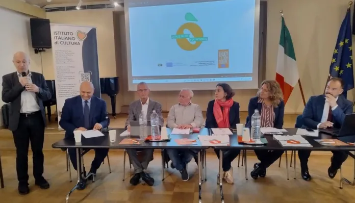Un relatore parla al microfono accanto a un tavolo con sei persone sedute; sullo sfondo si vedono uno schermo, la bandiera italiana e quella europea.