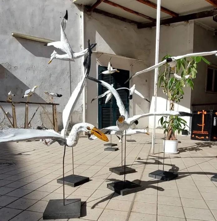 Sculture bianche a forma di uccelli, montate su supporti neri in uno spazio con pavimento piastrellato.