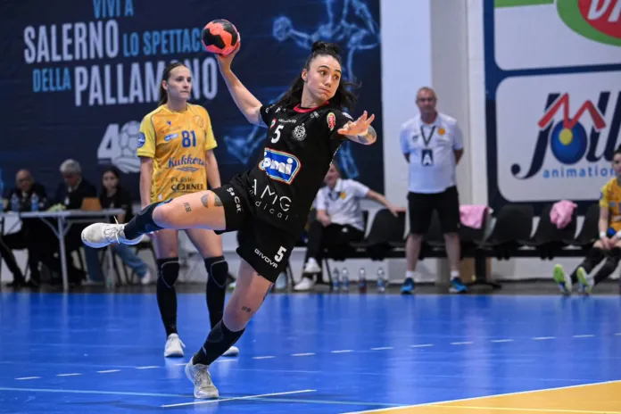 Una giocatrice in maglia nera salta e lancia la palla durante una partita di pallamano; sullo sfondo si vedono altre atlete e il campo.