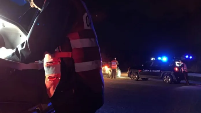 Un militare in uniforme è accanto a un’auto; sullo sfondo si vedono veicoli dei Carabinieri con i lampeggianti accesi.