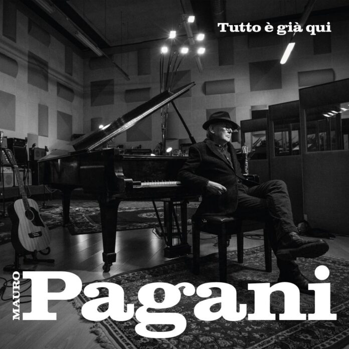 Foto in bianco e nero di Mauro Pagani seduto in studio di registrazione accanto a un pianoforte a coda, con chitarre in vista e atmosfera artistica.