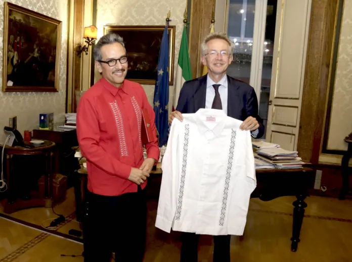 Due uomini in abiti formali posano accanto a un indumento bianco tenuto aperto.