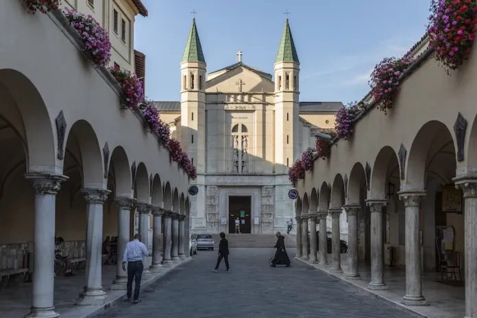 Facciata di una chiesa con due campanili, in fondo a un passaggio porticato decorato con fiori; alcune persone camminano nella piazza.