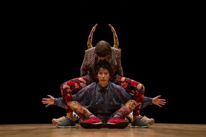 Tre danzatori sono disposti in una composizione simmetrica sul palco, con le braccia sollevate o distese; sullo sfondo nero e sul pavimento di legno.