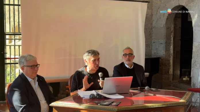 Tre persone siedono a un tavolo; quella al centro parla davanti a un computer portatile e tiene un microfono.
