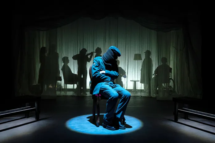 Una figura in abiti blu è seduta al centro di un palco illuminato, con sagome visibili dietro un fondale.