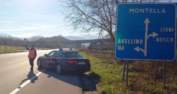 Un’auto dei Carabinieri è ferma sul margine della strada accanto a un agente; sul cartello stradale si legge «Montella».