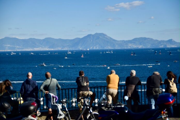Persone osservano le regate preliminari dell'America's Cup sul mare a Napoli con la vista delle montagne sullo sfondo.