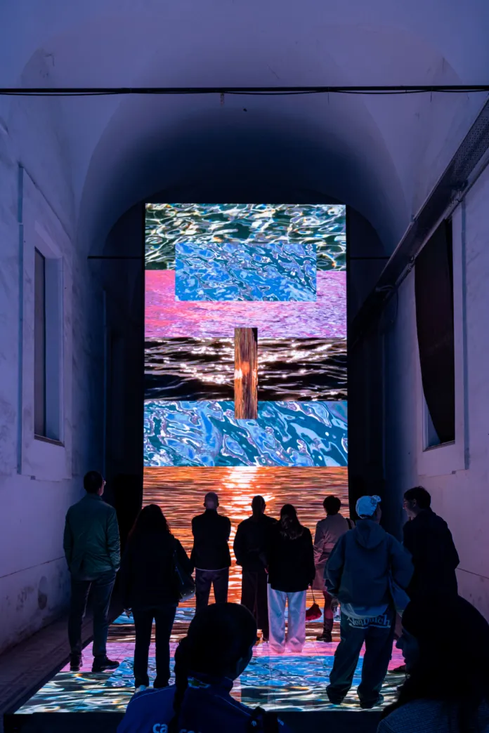 Persone che osservano un'installazione di videoarte luminosa e colorata in uno spazio interno con volta alta.