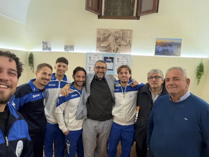 Un gruppo di persone posa in una stanza. Alcune indossano tute sportive blu e bianche.