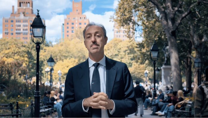 Maurizio Martina in abito formale presenta un discorso all'aperto con edifici e alberi sullo sfondo, durante una conferenza a New York.