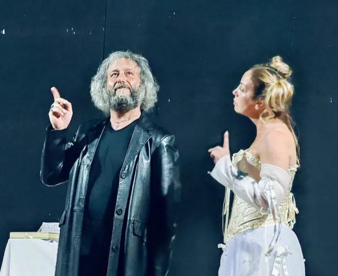 Due interpreti sono in scena davanti a uno sfondo scuro; uno dei due alza un dito mentre l’altra persona lo guarda.