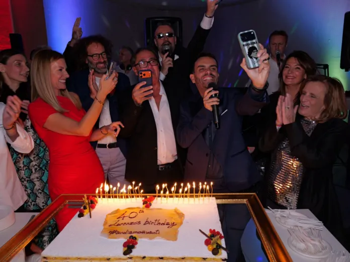 Persone festeggiano attorno a una torta con candeline; un uomo tiene un microfono e un’altra persona scatta una foto.