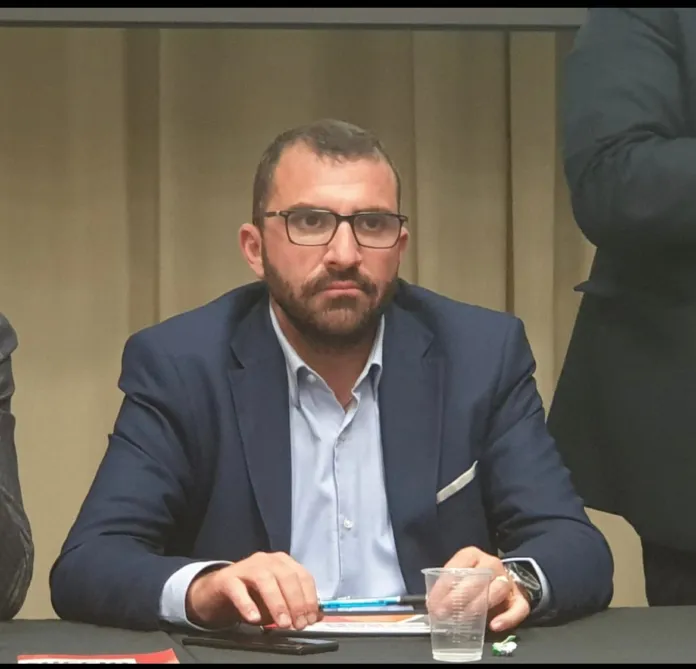 Un uomo con occhiali e giacca è seduto davanti a un tavolo con documenti e un bicchiere.