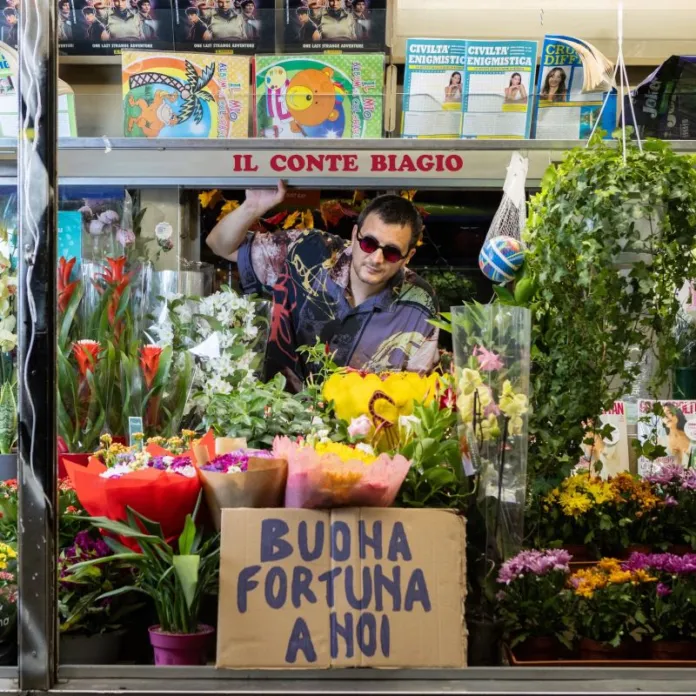 Una persona è visibile all’interno di un’edicola circondata da fiori; davanti c’è un cartello con la scritta «Buona fortuna a noi».