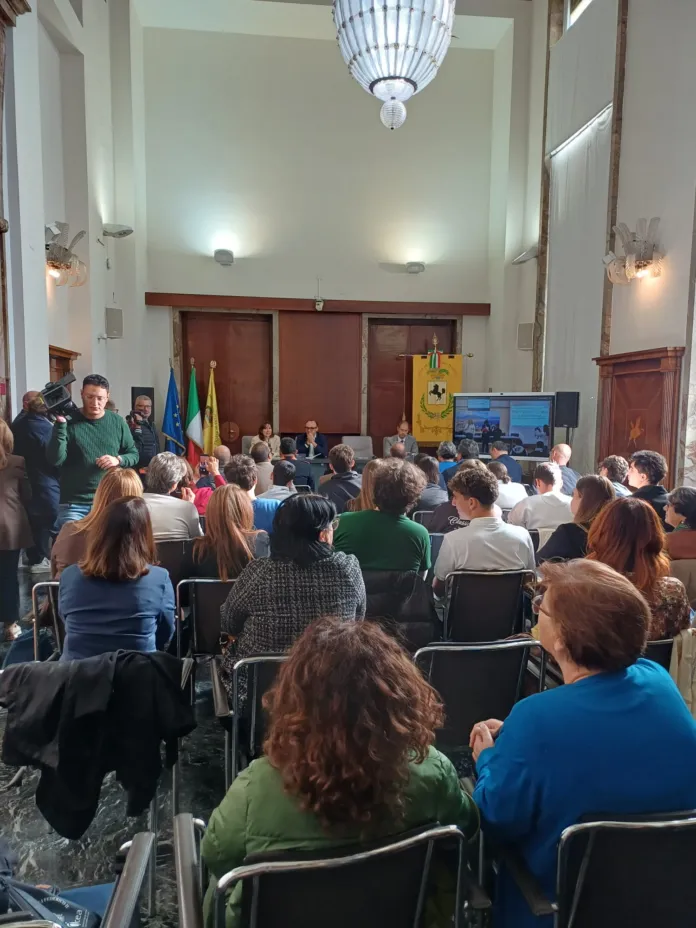 Persone sedute in una sala seguono una presentazione proiettata.