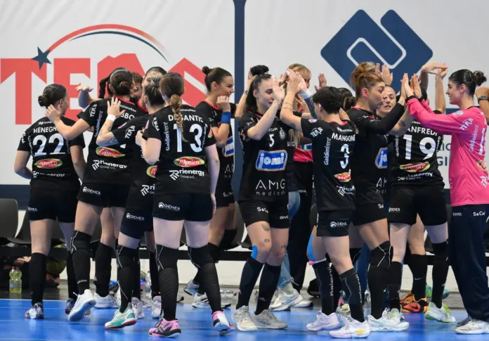 Un gruppo di atlete in divisa si raccoglie sul campo da pallamano; sullo sfondo si vedono cartelli pubblicitari.