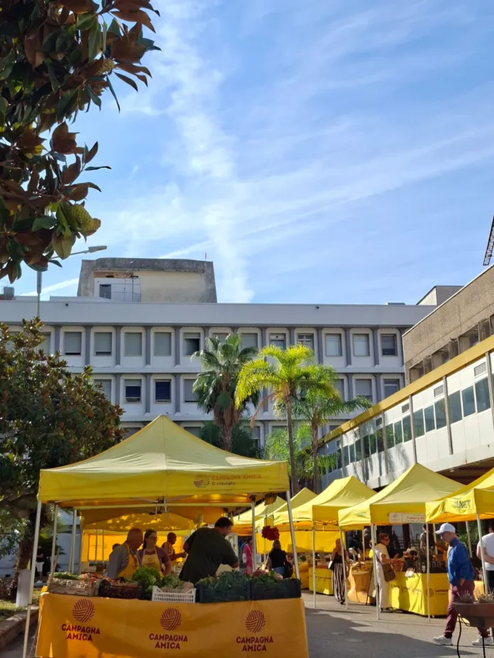 Stand di un mercato all'aperto con molte persone che acquistano ortofrutta, sotto tende gialle, in una giornata di sole.