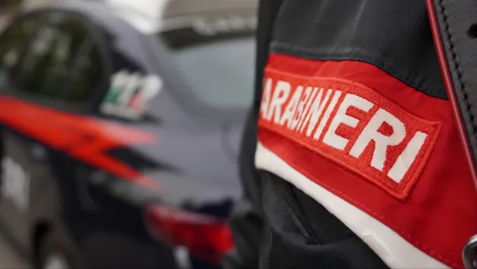 In primo piano una divisa con la scritta «Carabinieri»; sullo sfondo, sfocata, un’autovettura con bande rosse.
